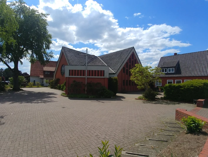 Rote Backsteinkirche im Licht der Sonne, umgeben von Bäumen, mit blauem Himmel und weißen Wolken.