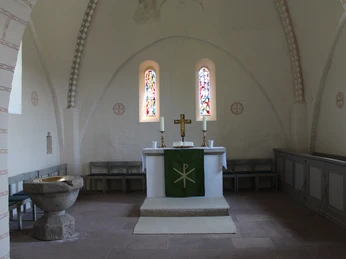 Ein lichter Kirchenraum mit Altar, Taufbecken und zwei farbenfrohen Glasfenstern im Hintergrund.
