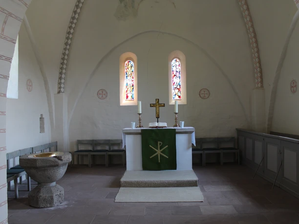 Ein lichter Kirchenraum mit Altar, Taufbecken und zwei farbenfrohen Glasfenstern im Hintergrund.