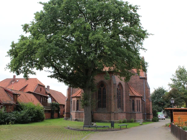 Backsteinkirche mit hohen gotischen Fenstern, umgeben von Bäumen und Fachwerkhäusern im Hintergrund.