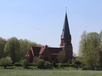Backsteinkirche mit hohem Turm in ländlicher Umgebung, umgeben von Bäumen unter blauem Himmel.