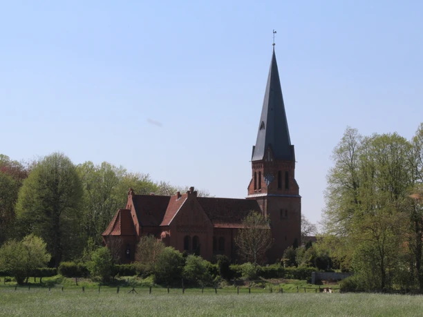 Backsteinkirche mit hohem Turm in ländlicher Umgebung, umgeben von Bäumen unter blauem Himmel.