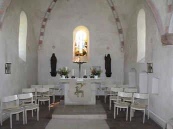 Ein lichtdurchfluteter Altarraum mit Kreuz, flankiert von Statuen, mit weißen Stühlen umringt.