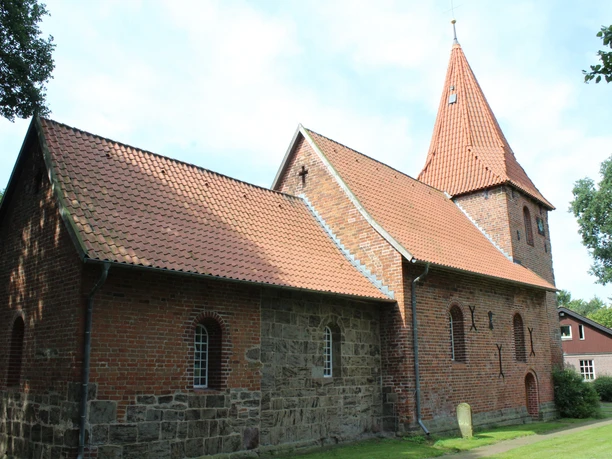 Backsteinkirche mit markantem Turm und rötlichen Dachziegeln, umgeben von grünen Bäumen und Mauerwerk.