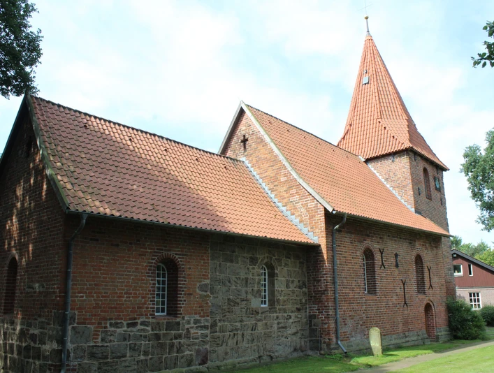 Backsteinkirche mit markantem Turm und rötlichen Dachziegeln, umgeben von grünen Bäumen und Mauerwerk.