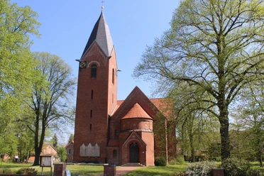 Backsteinkapelle mit spitzem Turm und Uhr, umgeben von Bäumen vor strahlend blauem Himmel.