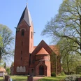 Kapelle Schweringen Backsteinkapelle mit spitzem Turm und Uhr, umgeben von Bäumen vor strahlend blauem Himmel.