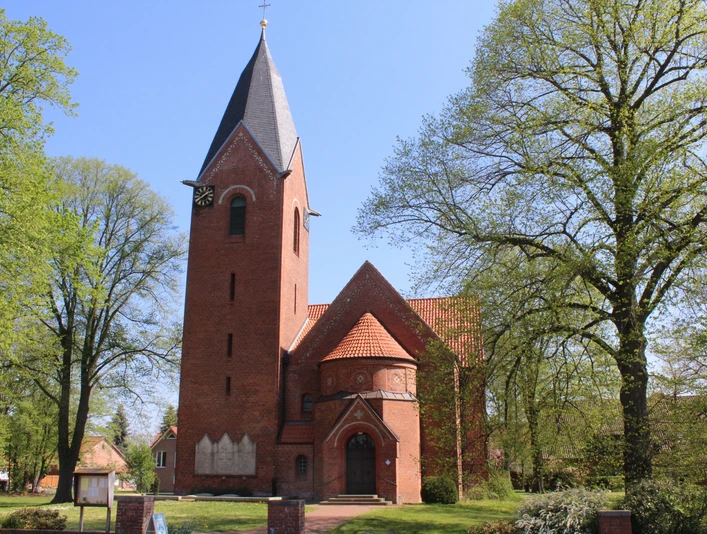 Kapelle Schweringen Backsteinkapelle mit spitzem Turm und Uhr, umgeben von Bäumen vor strahlend blauem Himmel.