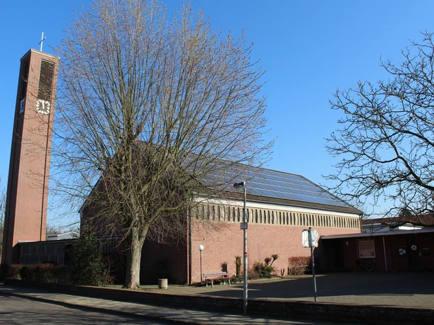 Backsteinkirche mit markantem Turm und Uhr, umgeben von kahlen Bäumen, an einem klaren, blauen Himmel.