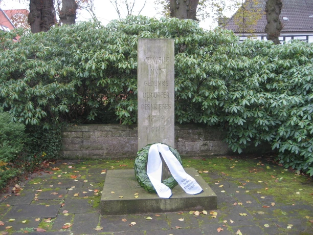 Steinernes Kriegerdenkmal mit Kranz in dichter Vegetation, gewidmet den Opfern des Krieges 1939–1945.