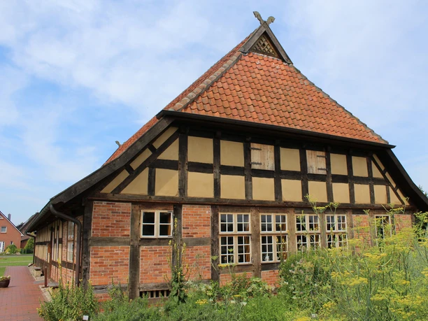 Fachwerkhaus mit roten Ziegeln und steilem Dach vor blauem Himmel, umgeben von grünem Garten.