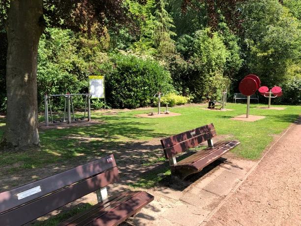 Grüne Parklandschaft mit verschiedenen Outdoor-Fitnessgeräten und Holzsitzbänken unter schattigen Bäumen.