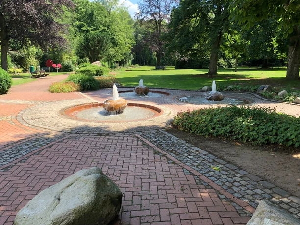 Ein geöffnetes Parkgelände zeigt einen Brunnen mit sprudelndem Wasser, umgeben von grüner Vegetation.