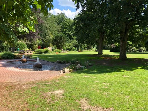 Ein gepflegter Kurpark mit sprudelndem Brunnen, üppigen Bäumen und weitläufigen Rasenflächen.