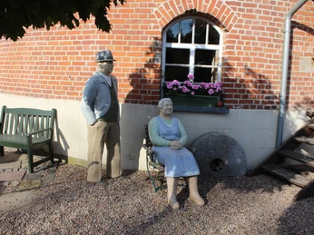 Windmühle Lahde Zwei Menschenfiguren, die vor einer roten Backsteinmauer auf einem kiesbedeckten Boden sitzen und stehen.