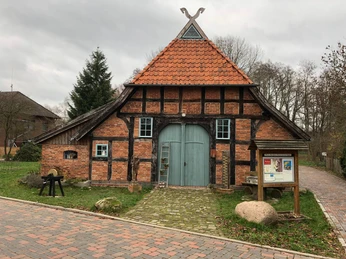 Häuslings Haus Historisches Fachwerkgebäude mit roten Ziegeln, blaugrauer Toreinfahrt, umgeben von Bäumen und Pflastersteinen.