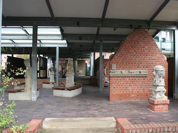 Lapidarium seitlich Ein überdachter Bereich mit Ziegelmauern und Skulpturen. Metallträger stützen die Holzkonstruktion.