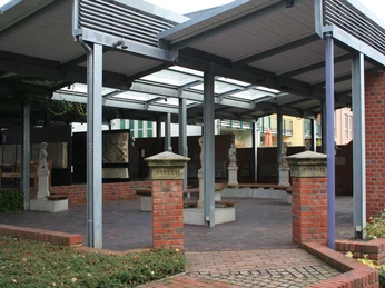 Ein überdachter Eingangsbereich mit roten Backsteinsäulen, Skulpturen und gläsernen Dachpaneelen.