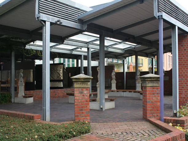 Ein überdachter Eingangsbereich mit roten Backsteinsäulen, Skulpturen und gläsernen Dachpaneelen.