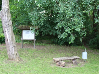 Landesbergen Lasseburg Grüne Umgebung mit einem hölzernen Sitzbereich neben einem Infokasten unter schattenspendenden Bäumen.