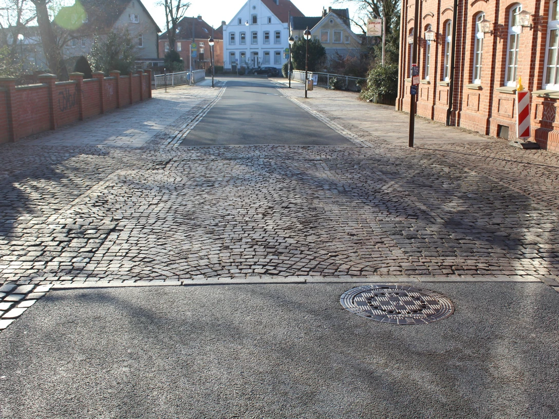 Straßenansicht mit Kopfsteinpflaster, flankiert von roten Backsteinhäusern und einem weißen Gebäude.