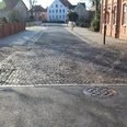 Straßenansicht mit Kopfsteinpflaster, flankiert von roten Backsteinhäusern und einem weißen Gebäude.