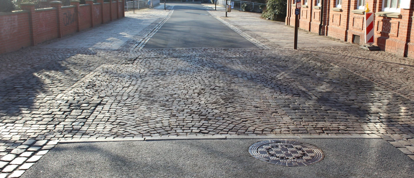 Straßenansicht mit Kopfsteinpflaster, flankiert von roten Backsteinhäusern und einem weißen Gebäude.