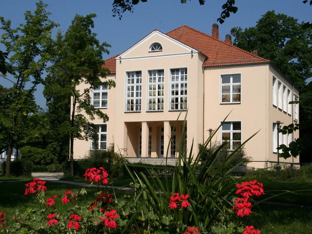 Kreismusikschule - Lindemann'sches Schloß Quadratisch-vornehmes Schlossgebäude mit roten Dachziegeln, umgeben von grünen Bäumen und roten Blumen.