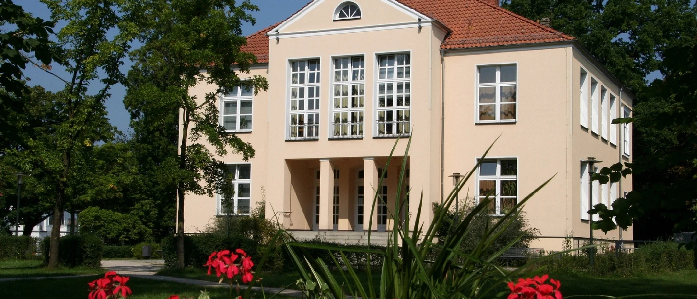 Kreismusikschule - Lindemann'sches Schloß Quadratisch-vornehmes Schlossgebäude mit roten Dachziegeln, umgeben von grünen Bäumen und roten Blumen.