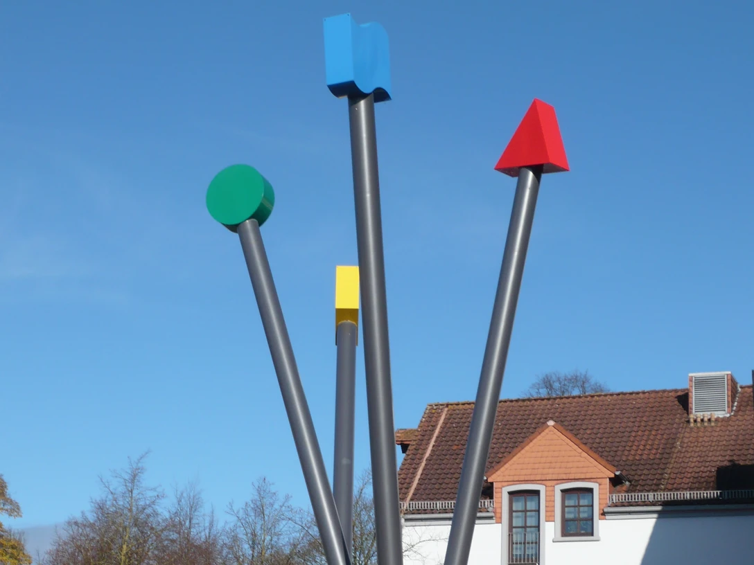 Logo-Stelen Vier unterschiedlich gefärbte, senkrechte Metallstangen als modernes Kunstwerk unter klarem Himmel.