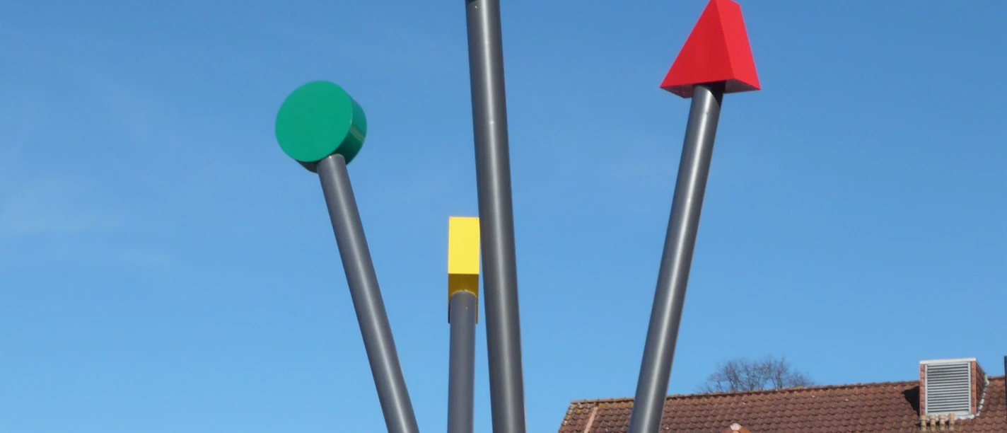 Logo-Stelen Vier unterschiedlich gefärbte, senkrechte Metallstangen als modernes Kunstwerk unter klarem Himmel.