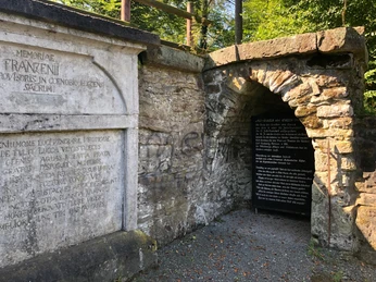 Luccaburg Historische Steinmauer mit lateinischer Inschrift und einem dunklen Eingang, umgeben von grünem Laub.