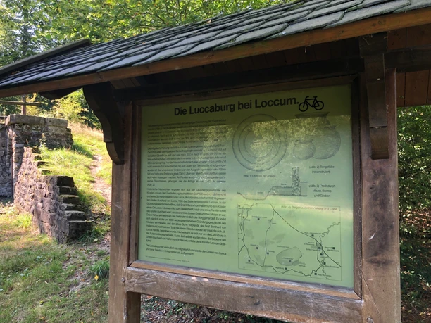 Information kiosk in a green space with text about Luccaburg near Loccum, next to it are old stone structures.