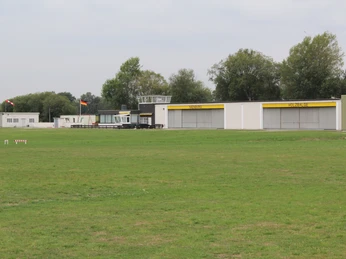 Grüne Wiese mit Hangars und Kontrollturm eines kleinen Flugplatzes unter bewölktem Himmel.