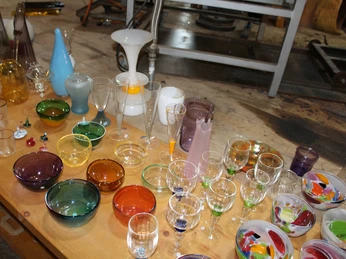 Gläser Glashütte Gernheim Verschiedene bunte Glaswaren, darunter Gläser, Schüsseln und Karaffen, liegen auf einem Holztisch.