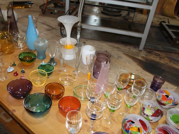 Gläser Glashütte Gernheim Verschiedene bunte Glaswaren, darunter Gläser, Schüsseln und Karaffen, liegen auf einem Holztisch.