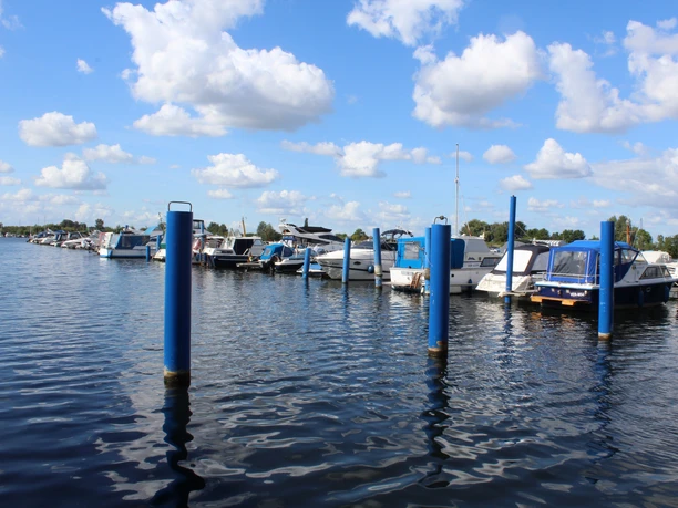 Boote liegen an blauen Stegen vor Anker, mit Wolken am Himmel und ruhigem Wasser darunter.