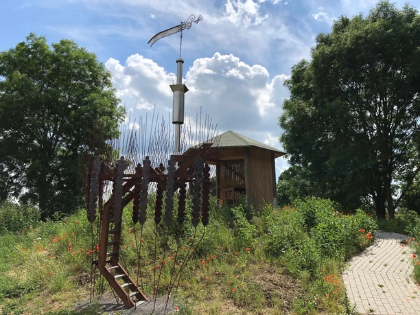 Marschbodenglück – Schibboleth Skulptur eines Widders aus Stahl, umgeben von Wildblumen und einem hölzernen Pavillon im Marschland.