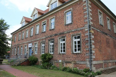 Historisches Backsteingebäude mit weißen Sprossenfenstern und Treppe, umgeben von Grünflächen.