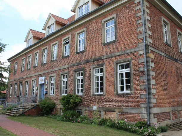 Historisches Backsteingebäude mit weißen Sprossenfenstern und Treppe, umgeben von Grünflächen.