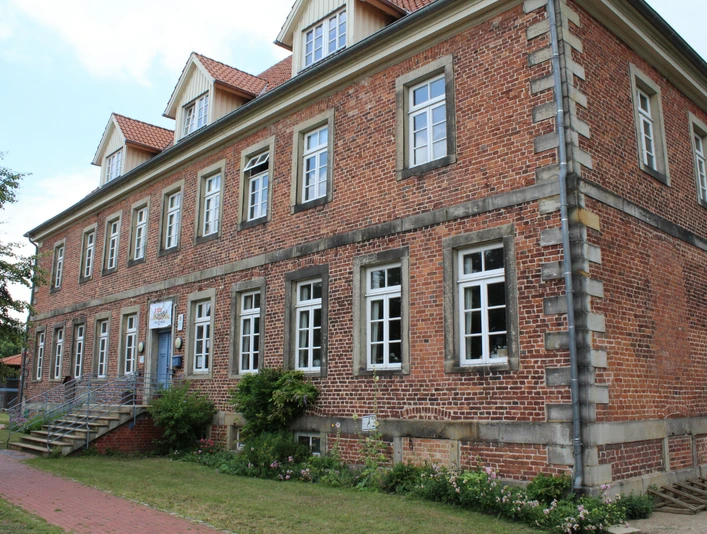 Historisches Backsteingebäude mit weißen Sprossenfenstern und Treppe, umgeben von Grünflächen.