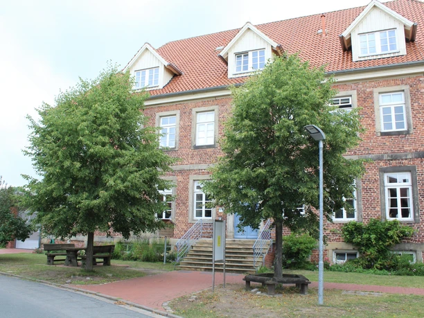 Ein Ziegelbau mit rotem Dach, Treppenzugang und Sitzbänken, umgeben von Bäumen und Strauchwerk.