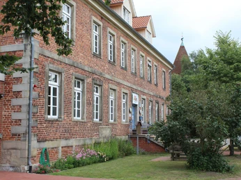 Mehrgenerationenhaus Ein großzügiges Backsteingebäude mit weißen Fensterrahmen, umgeben von grüner Natur und Blumen.