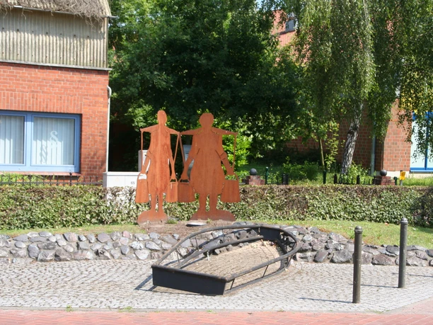 Zwei rostfarbene Skulpturen von landwirtschaftlichen Arbeiterinnen stehen vor einem roten Ziegelhaus.