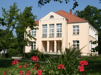 Ein zweistöckiges Schloss mit heller Fassade und großen Fenstern, umgeben von üppigem Grün.