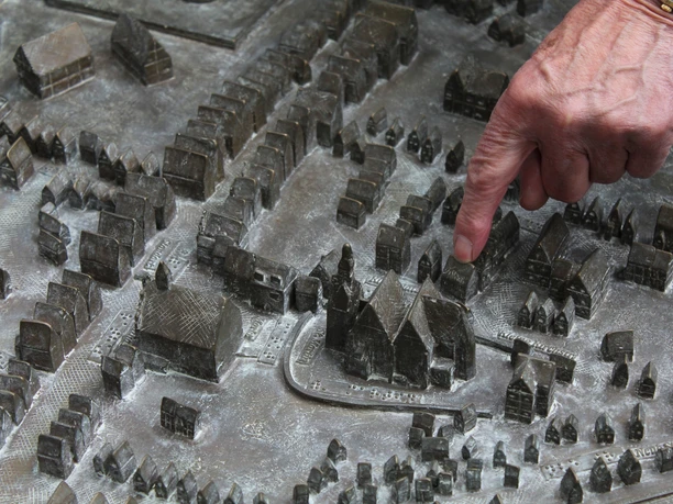 Stadtmodell Ein Finger deutet auf ein detailliertes Stadtmodell aus Bronze, das Gebäude und Straßen darstellt.