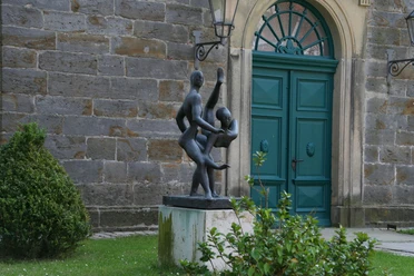 Zwei tanzende Figuren aus Bronze vor einem historischen Gebäude mit grünem Holztor und Pflanzen.