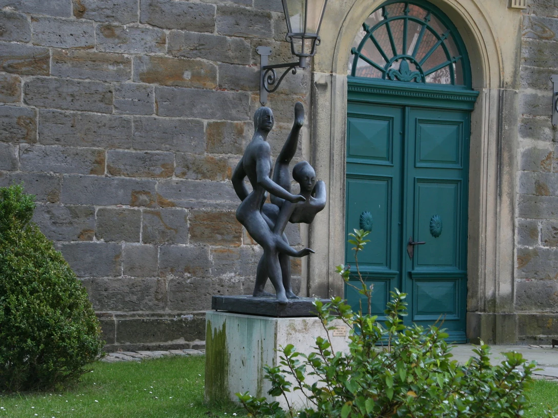 Zwei tanzende Figuren aus Bronze vor einem historischen Gebäude mit grünem Holztor und Pflanzen.