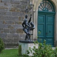 Zwei tanzende Figuren aus Bronze vor einem historischen Gebäude mit grünem Holztor und Pflanzen.