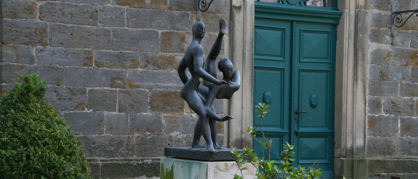 Skulptur "Paartanz" Zwei tanzende Figuren aus Bronze vor einem historischen Gebäude mit grünem Holztor und Pflanzen.